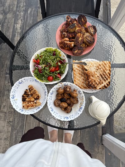 The image depicts an overhead view of a table set for a meal outdoors. The table is round with a glass top and a dark metal frame, set on a wooden deck. On the table, there are several dishes of food: a pink plate with grilled chicken and potatoes, a white bowl filled with a salad of greens, cherry tomatoes, and red peppers, a white bowl with grilled mushrooms, a white bowl with grilled chicken pieces, and two slices of grilled bread on a plate. A white pitcher sits near the grilled mushrooms. In the foreground, the legs and feet of a person wearing white slippers and a dark brown sock are visible, along with the lower part of a white robe or jacket. The background shows a wooden deck with some railing and a hint of sand or ground beyond. The overall impression is a casual outdoor meal. The specific location of Ryzhkovo, Russia is not visually evident from the image.