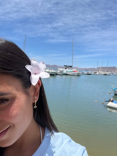The image shows a close-up of a woman's face. She has long, dark hair, and a white flower is tucked behind her ear. She is wearing a light blue shirt, a gold earring with a small charm that looks like an eye, and another small gold earring. She is smiling slightly. In the background, there is a body of water with multiple sailboats and small motorboats docked. The water is calm and blue, and the sky is clear and bright blue with some white clouds. The photo appears to be taken in a marina in La Paz, Mexico, with the coordinates 24.156, -110.324.  The image is likely taken from a slightly elevated perspective, as the water is visible in the background. The woman's face is in focus, while the background is slightly out of focus. The image is well-lit and the colors are vibrant.