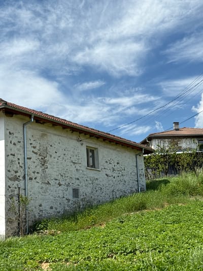 The image shows a stone house in Ogradna, Bulgaria, with a red-tiled roof. A window is visible on the stone wall. Tall green grass is growing along the side of the house. Another building with a similar roof is located behind it. Overhead, the sky is blue with wispy white clouds and power lines.
