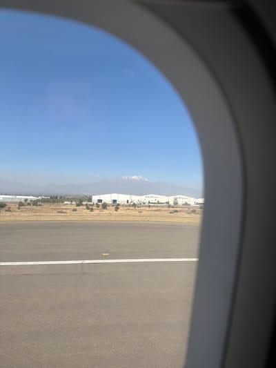 The image depicts an exterior view from an aircraft window, partially framed by the grey interior window structure, looking out over an airport environment near Santa María Zacatepec, Mexico, under a clear, light blue sky. In the immediate foreground, a dark asphalt runway or tarmac is visible, featuring a distinct white painted line running horizontally across its surface. A small yellow marker is present on the asphalt to the right of the white line.
Beyond the runway, a stretch of dry, yellowish-brown grass and scrubland extends, interspersed with sparse green foliage and small trees. In the midground, a series of large, white, low-rise industrial buildings are prominently featured. At least two of these buildings display the text "Multi Aceros" on their facades, indicating their function or proprietorship. Several indistinct vehicles, likely commercial or service trucks, are parked or moving near these structures.
In the distant background, a hazy mountain range stretches across the horizon. A distinct, taller mountain peak, capped with snow, rises prominently above the other mountains on the right side of the frame, consistent with Pico de Orizaba. No individuals are visible within the scene. The overall lighting suggests bright daylight conditions.