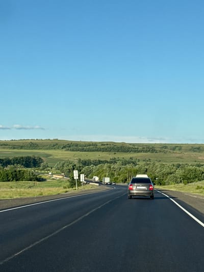 The image captures a road scene with a clear blue sky overhead and rolling green hills in the background. The road, paved with dark asphalt, curves gently to the left and has white lane markings. In the foreground, a dark-colored sedan is traveling on the road. Further ahead, on the left side of the road, there are several trucks and signs visible. The environment appears to be rural, with lush vegetation and trees bordering the road and hills. The location is identified as the city of Kendektamak, Russia.
