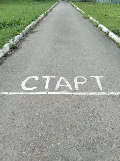 The image shows a paved asphalt path with a white line marking the start of a race. The word "СТАРТ" (START) is written in white chalk above the line. The path is bordered on both sides by green grass and a concrete curb. The scene appears to be outdoors in a park or recreational area. The location is specified as the city of Akberdino, Russia.