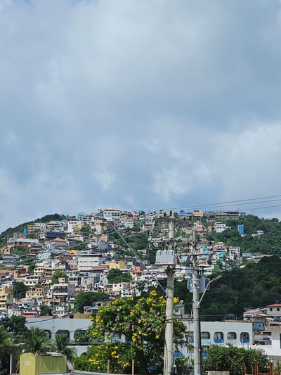 An elevated view captures a densely populated hillside in Arraial do Cabo, Brazil, under an overcast sky. The upper half of the image features a light grey, cloudy sky. Below, a steep, green-vegetated hillside is extensively covered with a multitude of closely packed, multi-story residential structures. These buildings, constructed from concrete and brick, display varied architectural styles and a palette of colors including white, beige, light blue, and grey, often with visible flat or tiled rooftops. Electrical infrastructure dominates the mid-ground, with a prominent concrete utility pole equipped with grey transformers, insulators, and numerous crisscrossing overhead power lines and communication cables. To the immediate left of the pole, a tree with clusters of bright yellow flowers is visible. Further left and lower, a segment of a light green, possibly corrugated metal, structure is present. In the lower mid-ground, a white building with several arched windows framed in blue is partially visible. No individuals are discernible within the frame.