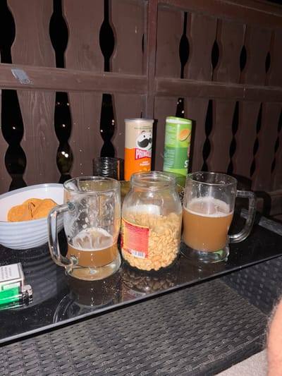 On a dark, outdoor table, likely a balcony or patio in Sohren, Germany, are several items suggesting a casual gathering. Two glass mugs of amber-colored liquid, possibly beer, sit on the table, one with a frothy head. Next to them is a large glass jar filled with small, light-brown nuts, likely peanuts. To the left, a white bowl contains several golden-brown potato chips. In the background, two cylindrical containers, one orange and white labeled "Pringles," and another green and white with an orange illustration, stand upright. A dark, ribbed object, possibly a glass ashtray or small container, is positioned between the Pringles cans. In the foreground, a pack of cigarettes and a green lighter rest on the table surface. The scene is set against a backdrop of dark wooden railing with an abstract pattern.
