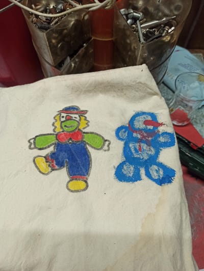 The image shows a close-up of a piece of light-colored fabric, possibly a tote bag or a dishtowel, adorned with two hand-drawn cartoon characters. On the left is a clown with a green face, a red and blue hat, yellow hair, and a red bow tie. It wears a blue jumpsuit and yellow shoes. To the right is a blue, teddy bear-like character with red accents on its head and chest. The fabric appears somewhat textured and has a slight stain in the lower right quadrant. In the blurred background, a metal container filled with screws, bolts, and other hardware is visible. A clear glass with red liquid and some red paper or cardboard are also present. The image does not provide any information or context that would allow for its location to be identified within the city of Werl, Germany.