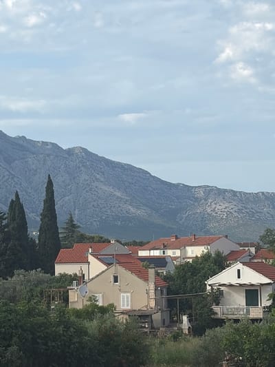 The image captures a residential landscape in Grad Korčula, Croatia, set against a backdrop of mountains under an overcast sky. The upper third of the image shows a pale blue sky with scattered stratocumulus clouds. Below the sky, a large, rugged mountain range, appearing greyish-blue due to distance and rock type, extends across the entire width of the frame, displaying visible erosion and sparse vegetation.
In the mid-ground and foreground, a collection of residential buildings with terracotta-tiled roofs are nestled among dense green foliage. Architectural details include light-colored stucco walls, predominantly beige and white, with multiple windows, some featuring external white louvered shutters. Prominent features on the houses include several chimneys. A satellite dish is mounted on the facade of one house in the mid-left, and solar panels are visible on the roof of another house slightly to the right of the center. A white-railed balcony extends from a white-walled house in the lower right. Tall, dark green cypress trees punctuate the left side of the scene, standing against the mountain range. In the lower foreground, partially obscured by various green trees and shrubs, an outdoor stone barbecue structure is visible next to a smaller, single-story building. No individuals or active elements are present in the scene. The lighting is diffused, indicating an absence of direct sunlight.
