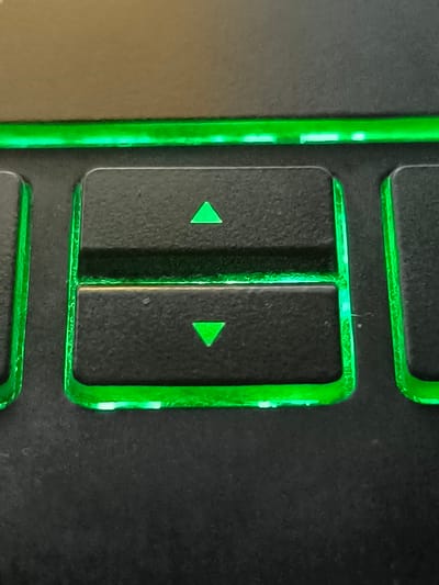 This media file is a close-up shot of a gaming keyboard, specifically focusing on the up and down arrow keys. The keys are black with illuminated green outlines and green arrows. The top key displays an upward-pointing green triangle, and the bottom key shows a downward-pointing green triangle. The keyboard appears to be in a dimly lit environment, with the green backlighting being the primary light source in the immediate vicinity. The texture of the keycaps and the surrounding keyboard surface is visible, showing a slightly rough, matte finish. There is no visible text on the keys themselves, beyond the directional arrows. The overall scene suggests a focus on the tactile and visual elements of a piece of computer hardware, likely used for gaming or other computer-intensive tasks. The location context provided, Makho, Georgia, does not offer any specific visual cues within the image itself, as the image is a close-up of an object.
