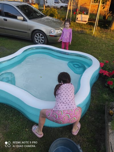 In Mliiv, Ukraine, two young girls are enjoying a sunny day in a backyard setting. One girl, wearing a pink long-sleeved shirt with a flower design and pink pants, stands near a parked car. The other girl, dressed in a pink shirt with bird motifs and pink shorts featuring rainbow patterns, is lying face down in an inflatable swimming pool filled with clear water. She appears to be reaching into the water with her head submerged. The scene suggests a warm, pleasant day, likely in the late afternoon given the golden sunlight casting shadows. Red geraniums bloom in the background, adding a touch of color to the grassy yard. Behind the standing girl, a fence and some wooden structures are visible, hinting at a residential area. The overall mood is one of playful outdoor activity and childhood enjoyment. The bottom right corner of the image contains visible text: "REDMI NOTE 9 PRO AI QUAD CAMERA".