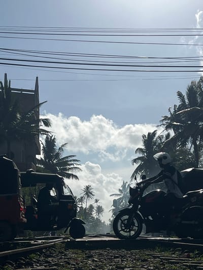 The image depicts an outdoor scene with a bright, cloudy sky and palm trees, suggesting a tropical or subtropical location. In the foreground, there are several power lines running vertically. To the right, a rough-textured surface, possibly a wall or embankment covered in stones and vegetation, is visible.
The central focus includes several vehicles. One is a motorcycle with a rider wearing a helmet. The motorcycle appears to be black and red, with a Yamaha logo visible on the wheel rim. Adjacent to it is a three-wheeled vehicle, a tuk-tuk, also dark in color. The tuk-tuk has a windshield and what appears to be a light on its roof. The vehicles are positioned on what looks like a road or pathway.
In the background, there are numerous palm trees, their fronds silhouetted against the sky. A partially constructed building with a roof structure is also visible amongst the trees, suggesting ongoing development or a rural setting.
The lighting indicates it is daytime, with the sun casting some glare and highlighting details, particularly on the power lines. The weather appears to be partly cloudy with bright sunshine. There are no overt emotional cues, but the scene suggests a typical day in a warm climate with vehicles and infrastructure. The overall impression is one of a busy, possibly tropical, environment. There is no visible text other than the "Yamaha" logo on the motorcycle.