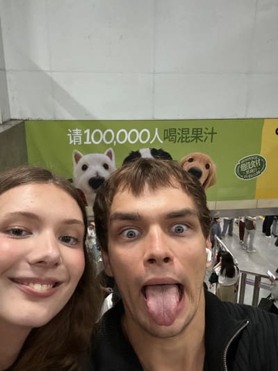 This is a selfie of a young couple in front of a large green banner in Xuhui District, China. The banner features an advertisement with cartoon dogs and Chinese text. The couple is making playful faces for the camera. The man has his eyes wide open and his tongue sticking out, while the woman is smiling broadly. The setting appears to be indoors, possibly a shopping mall or a public space, given the presence of other people in the background. The lighting suggests it is daytime. The banner contains Chinese characters, and the word "JUICE" is visible in English on a smaller circular logo. The prominent Chinese text on the banner appears to read "请100,000人喝混合果汁" which translates to "Inviting 100,000 people to drink mixed fruit juice."