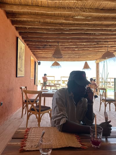 A person is seated at a table at a restaurant in Sidi Badhaj, Morocco. The person is wearing a striped shirt and a cap with a red star. They are holding a fork and a knife. A glass of red juice is on the table. A straw is in the glass. The restaurant has a thatched roof and orange walls. There are other people visible in the background. The table is set with woven placemats. A glass is empty on a place setting.