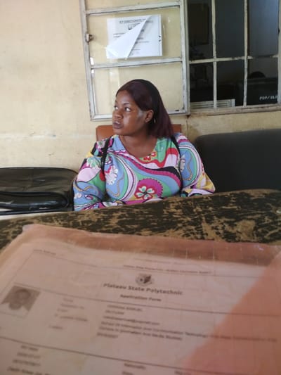 This media file shows a medium shot of a Black woman sitting at a desk in what appears to be an office or administrative building. She is wearing a colorful, patterned shirt and has dark hair. The woman is looking to her left, away from the camera, with a neutral expression.
The desk in the foreground is covered with a dark, textured fabric. On the desk, a partially visible document with a photograph of a person is laid out. The text on this document appears to be an application form for "Plateau State Polytechnic" and includes fields like "Name", "Application Form", and "School Of Information And Communication Technology And Mass Communication".
Behind the woman, a window with multiple panes is visible. One pane has a sign that reads "ICT DIRECTORATE OF". Another pane reveals a glimpse of the room behind, with some electronic equipment and a sign that says "PSP/BLD". The walls of the room are painted a light, creamy color.
The overall impression is that of an indoor setting, likely an educational institution in Bisichi, Nigeria, where the woman is possibly interacting with an administrative process or waiting to do so. The lighting suggests it is daytime. There are no strong indicators of weather conditions.