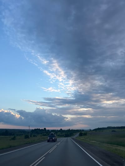 The image captures a scenic view of a road with a car driving on it, set against a backdrop of a dramatic sky. The perspective is from a vehicle on the same road, looking ahead. A dark-colored SUV is visible driving in the same direction, occupying the right lane of the asphalt road. The road itself is well-maintained, with clear white lane markings.

To the left of the road, a vast expanse of sky dominates the upper portion of the frame. Wispy, elongated clouds stretch across the blue sky, with some areas appearing darker and more ominous, suggesting changing weather. Sunlight peeks through some of the clouds, creating highlights and a sense of depth. The lower left portion of the image shows a lighter blue sky with more scattered, feathery clouds.

Beyond the road on the right, a lush green landscape unfolds, characterized by rolling fields and dense patches of trees. A few scattered trees are also visible in the distance, and a utility pole stands on the left side of the road. The horizon line is low, emphasizing the expansive sky.

The time of day appears to be late afternoon or early evening, given the quality of the light and the soft hues in the sky. The overall mood is one of a journey through a tranquil yet dynamic natural setting. The location is identified as Бикбарда, Russia. There is no visible text in the image.