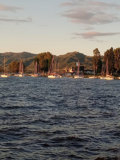 This image captures a serene lakeside scene, likely at either sunrise or sunset, in Villa Carlos Paz, Argentina. The foreground features a vast expanse of water, probably Lake San Roque, characterized by its textured surface with countless small ripples and reflections, indicating a calm body of water. Along the distant shoreline, numerous sailboats and other small boats are moored, their masts and rigging creating a dense, vertical pattern against the land. These boats are illuminated by a warm, golden light, suggesting the low angle of the sun. Behind the boats, a band of trees with dense, dark foliage lines the shore. In the background, rolling mountains rise, their earthy tones and rugged profiles silhouetted against the sky. The sky is filled with soft, pale hues, transitioning from a clear expanse to scattered, wispy clouds that are subtly touched with pink and orange, typical of dawn or dusk. The scene conveys a sense of peace and natural beauty. There is no visible text in the image.