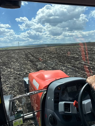 The image is a view from inside a Kubota tractor, possibly in a rural or agricultural area near Meshtitsa, Bulgaria. The steering wheel and dashboard of the tractor are prominent in the foreground. Through the windshield, a large field is visible, likely recently tilled or harvested, with rows of plant stubble. Beyond the field, the horizon shows a strip of greenery and possibly rolling hills. The sky is filled with clouds, some appearing dense and white against a blue background. The perspective suggests the tractor is in motion, possibly working in the field. A power line tower is visible in the distance to the left, further indicating a rural setting. The image is well-lit, with sunlight reflecting on the tractor's hood and dashboard. The label visible in the image is associated with Agritrade-ba.