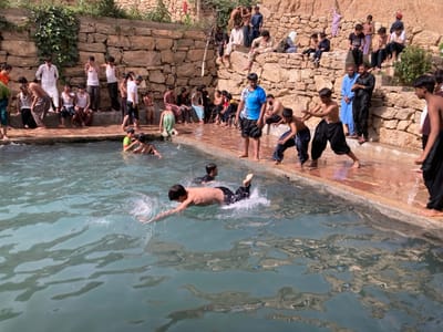 The image depicts an outdoor bathing and gathering scene in Killi Malik Sher Mohammad, Pakistan. A rectangular pool of light blue-green water occupies the foreground and midground, with multiple individuals swimming and standing at its edges. The pool is bordered by wet, light-colored concrete or tiled surfaces. Behind the pool, a multi-tiered retaining wall constructed of rough-hewn light brown stones rises, transitioning into a dry, earthen hillside in the background. Sparse green vegetation grows on top of and within the stone wall. The scene is brightly lit by natural sunlight, casting some reflections on the wet surfaces.
Approximately 40-50 individuals, predominantly males ranging from young children to adults, are present. Several women in traditional attire are visible on the upper terraces.
In the foreground, a shirtless boy in dark trousers is swimming on his stomach in the pool, creating splashes. Another boy, partially submerged, is visible further back. To the left, a person is seen mid-fall into the water, with only legs and lower torso visible above the water, generating significant splash. On the wet concrete edge to the right, two boys in dark trousers are engaged in a playful, wrestling-like interaction, one crouched and the other with arms raised. An adult male in a blue salwar kameez stands observing them. Another young male in a blue shirt and dark shorts stands near the water's edge, looking towards the pool.
On the various stone terraces behind the pool, numerous people are seated or standing, observing the activity below. Most are young males, some shirtless or wearing simple vests, others in traditional salwar kameez in various colors. A few individuals in the upper background are wearing more typical street clothes. The overall interaction suggests recreational activity and social gathering around the pool.