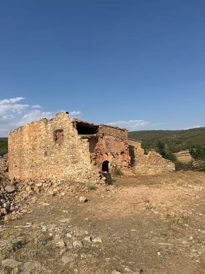 This is a vertical, outdoor, eye-level, medium shot of a ruined stone building under a clear blue sky with some scattered white clouds. The building appears to be old, with a partially collapsed roof and weathered stone walls. An arched doorway is visible on the side of the building. Dry, light brown grass and dirt cover the ground around the ruins. In the background, a lush green hillside rises, with trees and some cultivated terraces visible. The sun is shining, casting shadows, suggesting it is daytime, possibly late afternoon given the angle of the shadows. The scene evokes a sense of history and abandonment. There is no visible text in the image. The location context is Alpuente, Spain.