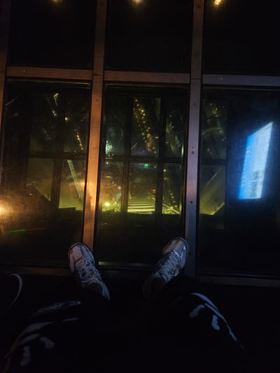 A high-angle view looks down through a glass floor, presumably high in a tower in Sumida, Japan. The glass floor is constructed in a grid pattern, with metallic framing around each glass panel. Through the glass, city lights and structures are visible, conveying the sense of height. A person is standing on the glass floor, as indicated by their shoes and legs visible on the right side of the frame. The scene suggests an observation deck or similar structure in a high-rise building within an urban environment.