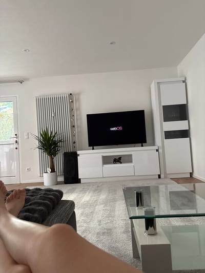 This is a medium shot, taken from a low angle, of a modern living room. The scene is brightly lit, suggesting it's daytime. Dominating the center of the frame is a large, black television screen displaying the "webOS" logo in pink. To the right of the TV is a white media console with drawers and shelves, one of which contains a small black figurine. Above the television, a white wall-mounted cabinet unit is visible, featuring both solid white doors and dark glass-fronted sections.
In front of the media console, a sleek, glass-top coffee table rests on a textured, light gray carpet. The coffee table has metallic cylindrical legs. To the left of the television, a white radiator is mounted on the wall. It is decorated with a string of small decorative stars and white spherical ornaments. A potted green plant with long, slender leaves sits in front of the radiator, in a white cylindrical pot.
On the far right of the image, the lower legs and feet of a person are visible, sitting on a dark gray sofa. The person's legs are crossed and bathed in light. A dark gray, faux-fur throw is draped over the sofa. Behind the sofa, a glimpse of the edge of the coffee table and the carpeted floor can be seen. A white door with glass panels and visible door handles is partially visible at the bottom of the frame, offering a view of greenery outside. The overall impression is one of a clean, minimalist, and comfortable living space in a German home.