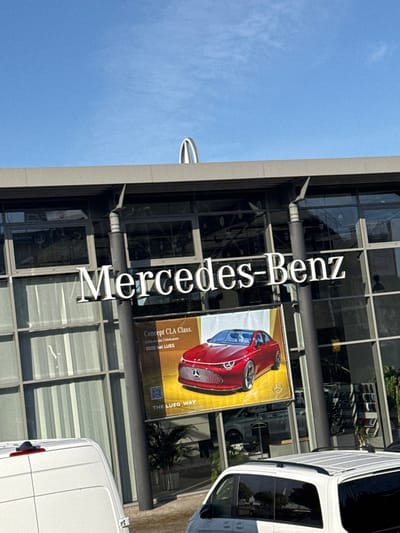 The image displays the exterior of a Mercedes-Benz dealership building under a clear blue sky in Hermsdorf, Germany. The modern building features a multi-story glass and dark grey facade. Large, white, illuminated letters spelling "Mercedes-Benz" are centrally affixed to the building's front. Above the main facade, a distinctive silver, arched architectural element is visible against the sky.

A prominent vertical banner hangs on the glass facade, featuring a red "Concept CLA Class" vehicle. Text on the banner reads "Concept CLA Class," "Entdecke das Unbekannte" (Discover the Unknown), "2025 bei LUEG" (2025 at LUEG), and "THE LUEG WAY," along with a QR code. Reflections of the sky and surrounding environment are visible in the glass panels.

In the foreground, the rear section of a white commercial van, possibly a panel van, is visible on the left, with a red light on its roof. To the right, the side and roof of another white passenger van or SUV, equipped with a roof rack, is partially visible. No individuals are discernible in the frame. The scene is illuminated by direct sunlight, indicating a clear weather day.