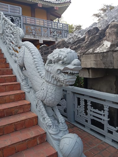 A detailed, medium, eye-level shot showcases a stone dragon statue along a staircase with red brick steps. The statue is grey-white and features a scaled body, intricate fur-like details, and a fierce expression with visible teeth. The dragon is positioned alongside a grey-white railing adorned with carved floral and geometric patterns. The dragon's tail extends up the staircase, while the head is situated near the railing's end, resting on a patterned spherical base.
In the background, a building with a yellow facade and a grey-white balcony is partially visible. Behind the railing, there is a rocky structure, possibly a man-made rock formation. The scene suggests the statue and staircase are part of a temple or historical site.
The location is Lumbini Sanskritik, Nepal, as specified in the prompt.