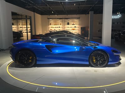 The image shows a blue McLaren Artura exhibited on a circular platform with a yellow LED border. The car is positioned inside a showroom; other cars and retail displays are visible in the background. A pillar bearing the McLaren logo is on the right side of the frame. The scene appears to be indoors in a showroom location and may be in Glattbrugg, Switzerland.