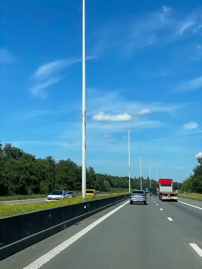 A bright, sunny day with a clear blue sky and scattered white clouds is depicted. The scene is a highway with a lane of grey asphalt and white lane markings. Trees line the right side of the highway, with a yellow verge featuring small yellow flowers. Several tall, white lampposts with visible rust streaks are spaced along the side of the road.

Traffic is visible on the highway. In the distance, a red and silver truck is traveling in the same direction as the viewer. Closer to the viewer, a silver car is in the left lane, and a white car is parked on the shoulder on the right side of the highway, appearing to be involved in an incident. A yellow vehicle, possibly a van, is also parked on the shoulder further down the road.

The image appears to be taken from a moving vehicle, as suggested by the slight blur and the perspective. The overall impression is one of a typical summer day on a European highway. There is no visible text in the image. The context provided indicates the location is Zandhoven, Belgium.