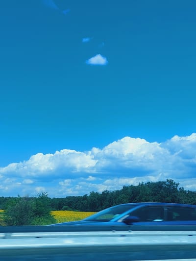The image captures a bright, sunny day with a vibrant blue sky and fluffy white clouds. In the foreground, a blue car is in motion, its side visible as it passes by. A light gray barrier runs horizontally across the bottom of the frame, suggesting the perspective is from a road or highway. Beyond the barrier and the car, a field of bright yellow flowers, likely sunflowers, stretches out, bordered by a dense line of dark green trees. The overall scene evokes a sense of travel and natural beauty, possibly on the outskirts of Malko Tranovo, Bulgaria, given the natural landscape.