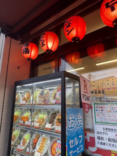 This is a vertical, eye-level shot taken outside a Japanese restaurant. The foreground features several bright orange paper lanterns hanging at different heights, some adorned with black Japanese characters. Behind the lanterns, through the restaurant's glass facade, a display case showcases an array of realistic food replicas, predominantly ramen and other Japanese dishes. Various payment method logos are visible on a sign in the upper right portion of the image, along with text indicating that photography and live streaming are not permitted inside the establishment. A "R POINT" banner is also visible. The overall lighting suggests it may be daytime or early evening, given the brightness and the reflections on the glass. The setting is a commercial street in Matsudo, Japan, evident from the Japanese text on signs and food displays.
