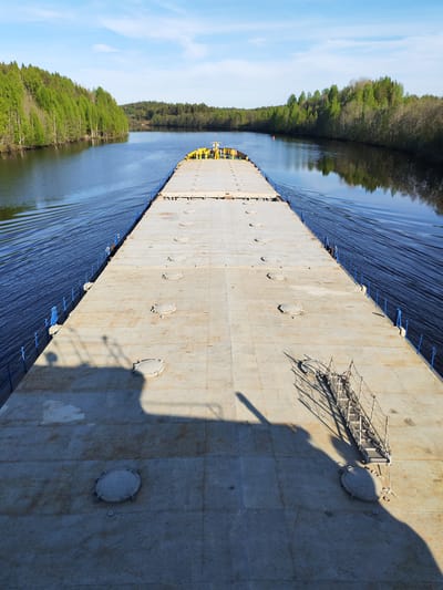 A high-angle shot shows a long, flat-topped barge sailing on a river in Depo, Russia. The barge, mostly empty, features a series of circular hatches and blue safety railings along its edges. At the far end of the barge are some yellow structures. The shadow of the photographer and a small structure is visible in the foreground. The river flows between green, forested banks, and the sky above is mostly clear with a few scattered clouds. The water is dark and reflective, with ripples indicating the barge's movement.