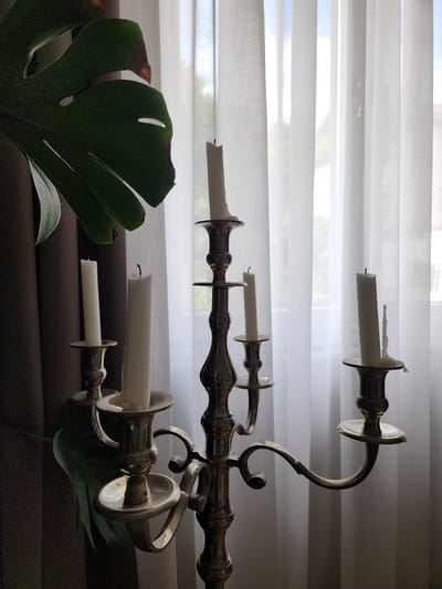 This image depicts an indoor still life, reportedly located in Idar-Oberstein, Germany.
The central subject is a large, ornate metallic candelabra, positioned slightly to the right of the frame's center. It appears to be made of polished brass or silver-plated material, featuring five branches holding white taper candles. All five candles are unlit. The candles exhibit varying degrees of prior burning, with visible wax drips down their sides and blackened wicks. The candle on the far right shows the most significant accumulation of melted wax.
To the left of the candelabra, a large, dark green, fenestrated leaf from a Monstera plant occupies the upper left portion of the image. The leaf displays characteristic splits and some visible browning along its edges. Behind the leaf and on the far left, a dark brown or black curtain is partially visible, creating a contrast with the brighter background.
The background consists of a window covered by translucent white curtains. These curtains are vertically pleated, allowing diffused natural light to enter the room. Faint, indistinct shapes suggestive of outdoor foliage and possibly distant buildings can be seen through the sheer fabric, indicating a daytime setting. The overall lighting is soft, with the foreground elements appearing somewhat in shadow against the brighter backdrop.