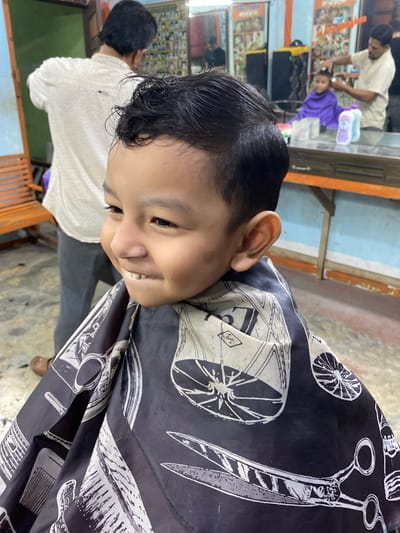 In a barbershop located in Payatieng, Indonesia, a young boy is getting a haircut. He is wearing a black cape with a pattern of scissors and other barber tools. The boy is smiling and looking to his left, with his teeth slightly showing. In the background, a barber is tending to another child, and a wall with framed pictures of hairstyles is visible. The environment appears to be a typical Indonesian barbershop, with wooden furniture and brightly colored walls.