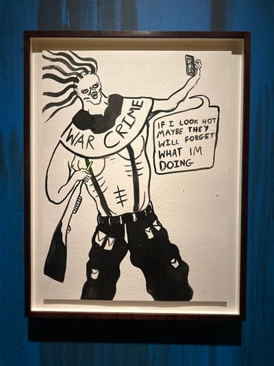 This image displays a framed ink drawing mounted on a textured, vertically streaked blue wall in the City of Westminster, United Kingdom. The drawing, executed in black ink on white paper, features a stylized, bare-chested figure with exaggerated features including spiky hair, a menacing facial expression, prominent ribs, and visible suspenders over its torso. The figure wears tattered black pants. A banner or collar encircles its neck, bearing the capitalized text "WAR CRIME." In its left hand, the figure holds a rifle vertically with the barrel pointing downwards, exhibiting a subtle green tint. Its right arm is raised, holding a smartphone as if taking a selfie. Adjacent to the figure's head, a speech bubble contains the text: "IF I LOOK HOT MAYBE THEY WILL FORGET WHAT I'M DOING." The artwork is enclosed within a dark brown wooden frame, offset by a white mat. The scene is illuminated, casting a distinct shadow directly beneath the frame on the blue wall.