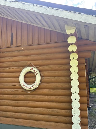 The image shows the exterior of a wooden building, likely a sauna, in Saue, Estonia. The building is constructed with vertical logs painted a warm brown color. A decorative element made of light-colored, round wooden pieces is attached along the top edge of the log wall. A white life preserver hangs on the wall, displaying red text that reads "MAJA SAUNAKULA". The life preserver is attached to the wall by a rope. The roofline is visible in the upper left, with wooden beams and some string lights. The scene suggests a sunny day, with some clouds in the sky. The overall impression is that of a rustic, outdoor recreational facility.