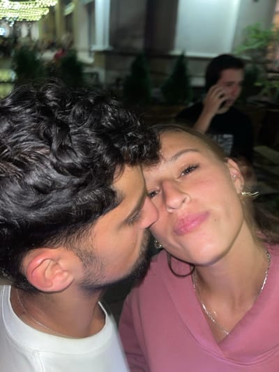 The scene is located in Arlington, United States, at night, outdoors. Two individuals are in the foreground, positioned intimately. On the left, a male with dark, curly hair and a short beard wears a white t-shirt and a thin silver chain. His face is angled towards the female, with his nose nearly touching hers. On the right, a female with light brown hair, wearing a pink hooded sweatshirt and a thin silver chain, looks directly at the camera with slightly puckered lips. She has a silver hoop earring and a distinct gold lightning bolt-shaped earring in her left ear. In the blurred background, a third male wearing a dark t-shirt is visible, holding a dark-colored mobile phone to his right ear, appearing to be conversing. The environment features out-of-focus building facades, several small, cone-shaped evergreen shrubs, and decorative string lights overhead. A faint 'P' sign is discernible in the upper left background.