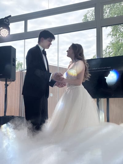 A young man in a tuxedo and a young woman in a white ballgown are holding hands. They are standing in front of a large window with trees visible outside. There is a screen to the right of the couple with Russian text on it. There is a speaker to the left of the couple. Smoke or fog is billowing around their feet. The text on the screen reads "signal is weak or absent. Check that the device is properly connected." The scene appears to be indoors, possibly at an event or ceremony in Osipovo, Russia.