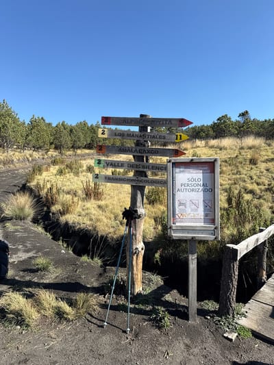 The image shows a signpost at what appears to be a mountain trail, likely in Tlamacas, Mexico, given the signs referencing Iztaccíhuatl-Popocatépetl National Park. The signpost displays trail names and distances denoted by numbers (2 and 11). A framed sign on the right reads "SÓLO PERSONAL AUTORIZADO," indicating a restricted area. Below this are pictograms prohibiting vehicles, bicycles, and littering. Trekking poles are leaning against the signpost. The environment includes grassy terrain and trees under a clear blue sky. The ground around the signpost appears to be bare and rocky.