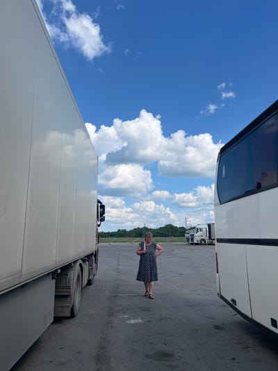 A middle-aged woman stands in an open parking lot next to a large white truck and a white bus under a partly cloudy blue sky. The woman is wearing a black and white polka dot dress and sandals. She is looking towards the ground with her hands on her hips. The truck is on the left side of the frame, and its side is mostly visible. The bus is on the right side of the frame, and its rear and side are visible. In the background, there are more trucks parked, along with trees and some wooden structures. The ground is asphalt. The overall scene suggests a rest stop or truck depot.