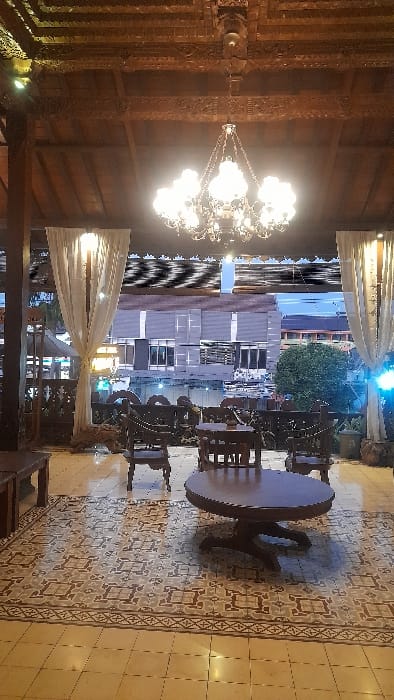 The image presents an unoccupied open-air or semi-enclosed seating area, identified as being in Glantengan, Indonesia. The overhead structure features a dark, ornately carved wooden ceiling with visible beams. A large, multi-lamped chandelier, illuminated and appearing metallic with glass globes, hangs centrally from the ceiling. The floor comprises a central section of intricate patterned tiles in shades of grey and brown, surrounded by plain, lighter-colored floor tiles.
The foreground contains a large, round, dark-wood table on a single pedestal base. Several dark-wood chairs with carved backrests and legs are arranged around or near this table and other smaller, rectangular dark-wood tables visible further into the space. A long, low wooden bench or table is partially visible on the left side of the frame. White, floor-length curtains are draped and tied back at the perimeter of the seating area. Beyond these curtains, a blurred urban background is discernible, featuring multi-story buildings with windows and a dark, reflective surface that appears to be water, exhibiting horizontal distortion artifacts. Green foliage is visible to the right, outside the immediate seating area. No individuals are present, and no specific actions or interactions are depicted.