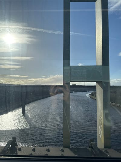 A partial view through a framed window reveals a sky with bright sunlight and wispy clouds on the left, contrasted by a body of water on the right. The water glistens in the sunlight, with a visible shoreline. Structural elements of a bridge or building frame the scene, with metal beams and supports visible. There are reflections in the glass, including hints of a person.