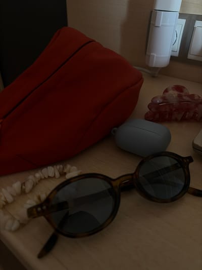 A close-up, medium shot is captured from a slightly elevated angle, looking down at a wooden surface. On the left, a bright orange pouch is partially visible, its fabric textured and its zipper prominently featured. To the right of the pouch, a light blue, oval-shaped object, possibly an earbud case, rests on the wood. Further to the right, a red and white marbled hair claw clip is positioned. In the foreground, a pair of tortoiseshell-patterned sunglasses with dark lenses are laid out, their arms resting on the surface. To the left of the sunglasses, a light-colored beaded necklace is casually placed, its irregular beads forming a curved line. In the background, a white electrical adapter is plugged into a wall socket. The lighting is dim and warm, casting soft shadows. There are no people visible in the image. The scene appears to be an indoor setting, possibly a bedside table or dresser, with no specific landmarks indicating Riudoms, Spain.