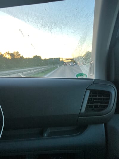 The image depicts a view from inside a vehicle, likely a car or van, traveling on a highway. The windshield is somewhat dirty, obscuring the view ahead. The scene outside shows a highway with multiple lanes and other vehicles in the distance. The light suggests it might be either sunrise or sunset. In the foreground, a portion of the vehicle's dashboard is visible, including an air vent. A green sticker is affixed to the windshield, indicating a valid emissions test required for vehicles. Oberschleißheim is not explicitly apparent in the image, but the highway setting and European registration sticker could suggest it is indeed in the region of Germany.