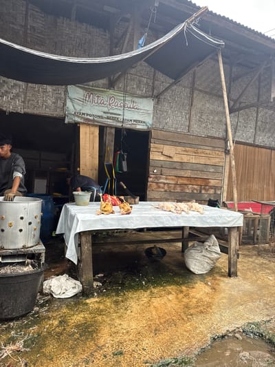 The image depicts an outdoor scene in Cotcut, Indonesia, likely a market stall. In the foreground, a table draped with a white cloth displays several prepared chickens. To the left, a young man in a grey shirt and gloves is working near a large metal poultry defeathering machine and a blue barrel. Behind the table, a second man in a dark shirt and blue apron is visible, leaning over and looking into a shelf. The stall is constructed with bamboo siding and a corrugated metal roof, with a banner overhead reading "Mita Rasuna" and indicating services for "AYAM POTONG - BEBEK - AYAM MERAH" (cut chicken, duck, red chicken). The ground is a mixture of dirt, concrete, and some standing water, with debris and scattered feathers suggesting the nature of the business. Various containers, a cage, and tools are also present.
