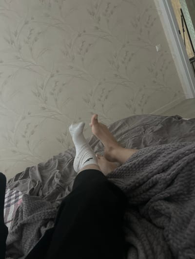 Two individuals are lying in bed in a room. The room has wallpaper with a subtle floral pattern. One individual's legs are visible, wearing a white sock and black pants. Another individual's legs are also visible, one foot bare and the other partially covered by a gray blanket. A gray blanket is draped over the bed. The scene appears to be indoors, likely in a residential setting in Veshki, Russia.