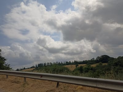 The image depicts a rural landscape with a guardrail in the foreground. The guardrail, made of metal, runs horizontally across the frame, appearing to be a safety barrier on the side of a road. Beyond the guardrail, the landscape unfolds into rolling hills covered with dry, golden grass. Further in the distance, there is a dense line of trees that forms a dark green horizon. Above the trees, a dramatic sky is filled with large, fluffy white and grey clouds, suggesting an overcast or potentially stormy day. The scene is consistent with a view from a road in a rural area, possibly during the summer months given the dry vegetation. There are no people or animals visible in the image. The location is identified as Digah, Azerbaijan.