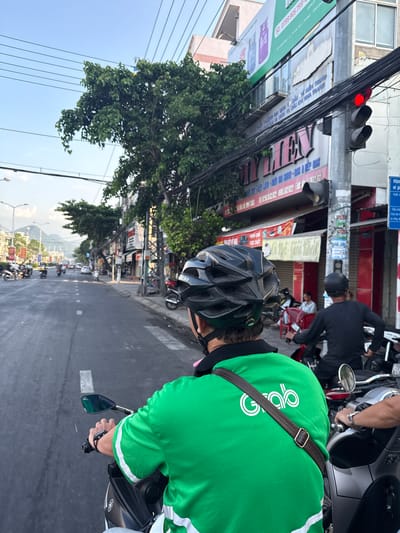 The image depicts a street scene in Bac Nha Trang Ward, Vietnam. In the foreground, a person wearing a bright green shirt with the "Grab" logo and a black helmet is riding a motorcycle. The rider's left hand is visible on the handlebars. To the right, another person, also wearing a black helmet, is riding a motorcycle. In the background, there are buildings with signage, trees, overhead power lines, and other vehicles on the road, including motorcycles and a car. The sky is clear with some clouds, and the scene appears to be during daylight hours. A traffic light with a red light illuminated is visible on a pole.
