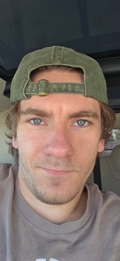 A close-up, head-on, eye-level selfie features a young white man wearing a green baseball cap and a tan t-shirt. He has light brown hair, fair skin with visible pores and some stubble on his face, and bright blue eyes. His eyes are looking directly at the camera, and he has a slight smile. The background is dark and out of focus, suggesting he is inside a vehicle, possibly with a sunroof or a dark interior. The lighting is bright, creating a natural look. The image was captured in Bremerton, United States of America.