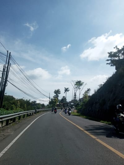 The image depicts an uphill, asphalt two-lane road curving left, under a bright blue sky with scattered white clouds, in Ban Phonhxavanh, Laos. On the far left, a cluster of dark utility poles supports numerous tangled black overhead cables, extending alongside a silver metal guardrail. Beyond the guardrail, dense green vegetation covers a sloping terrain. On the right side of the road, a steep, dark rock face or earthen embankment rises abruptly, topped with trees and sparse foliage. In the middle distance, a group of approximately five to seven individuals on dark motorcycles travels away from the viewer, ascending the curve. Another motorcycle, with a rider wearing a white helmet, is partially visible on the far right edge of the frame, also heading uphill. Sunlight casts shadows on the right side of the road. The road features a solid white line marking the far left edge, a broken white line for the lane division, and a solid yellow line on the right side of the visible lane.