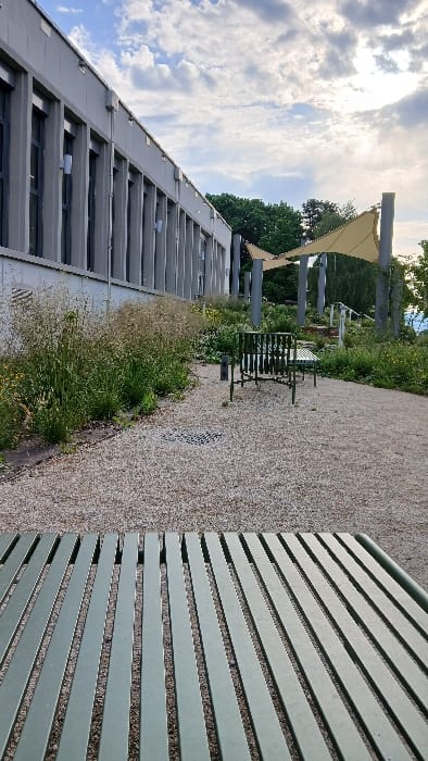The image shows a long, gray building with many windows, located in Besançon, France at coordinates 47.246, 5.987. In front of the building is a grassy area with a gravel path. There are two metal benches positioned along the path, with a third empty bench in the foreground. The metal benches are green in color and have a slatted design. The sky is partly cloudy, and a large, yellow, fabric canopy is visible in the background. Trees and foliage are also present, with the canopy partially shading the area. The image is taken from a low angle, looking towards the building and benches. There are no people present in the scene.
