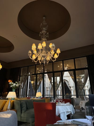 The image depicts the interior of a dining room in a restaurant or hotel located in Ouled Jelal, Morocco. The room has a high ceiling with a large, ornate chandelier hanging from the center. The chandelier is made of crystal and has many lights that illuminate the room. The room has large windows that look out onto a courtyard or garden. There are several tables set for dining, with white tablecloths and chairs. There are also several green, brown, and red upholstered chairs, some of which are visible in the foreground. The room has a sophisticated and elegant atmosphere.